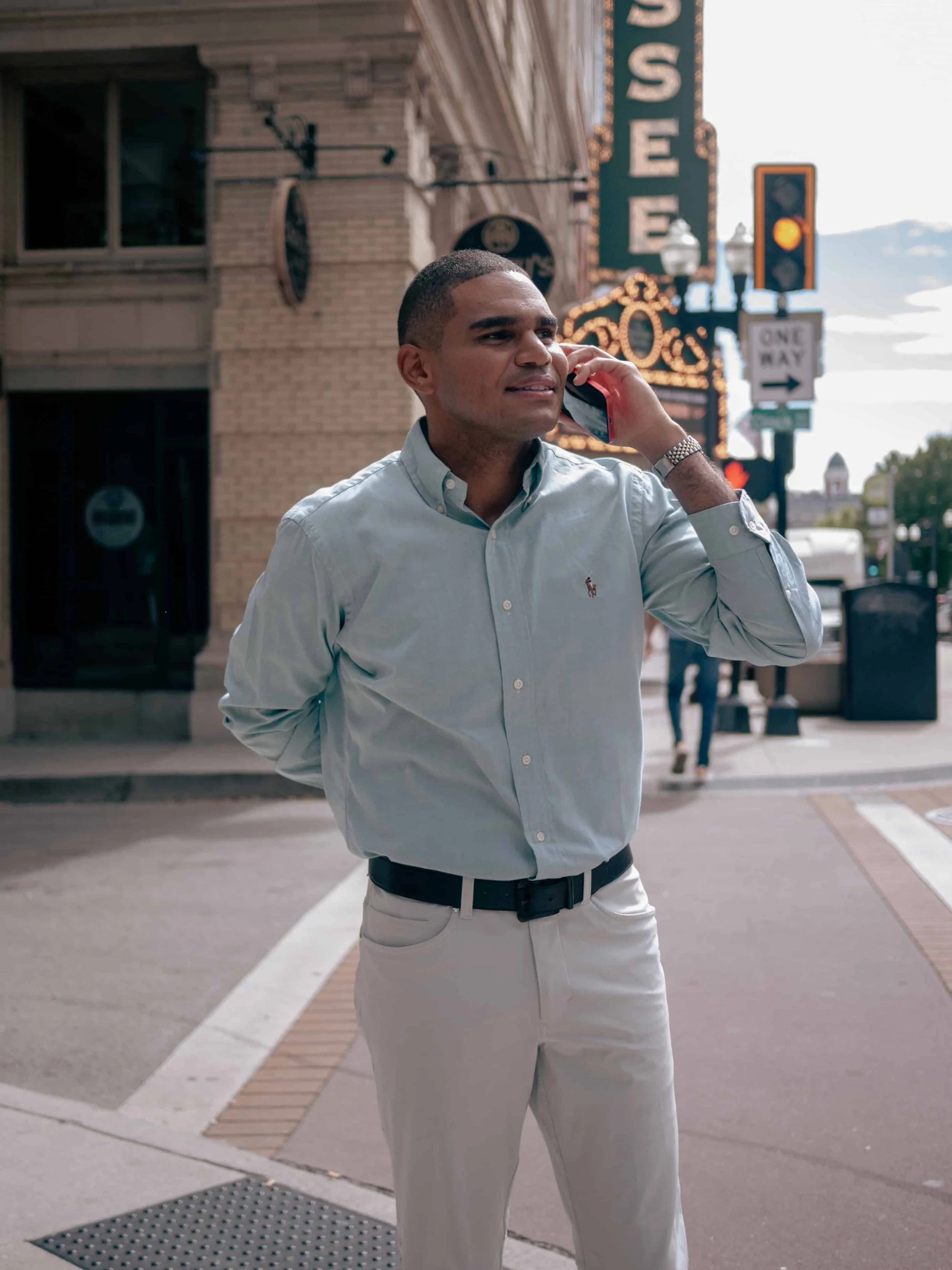Ty Briggs stands on a downtown sidewalk while talking on a cellphone, wearing a light blue button-down shirt and white pants with city buildings and street signs behind him.