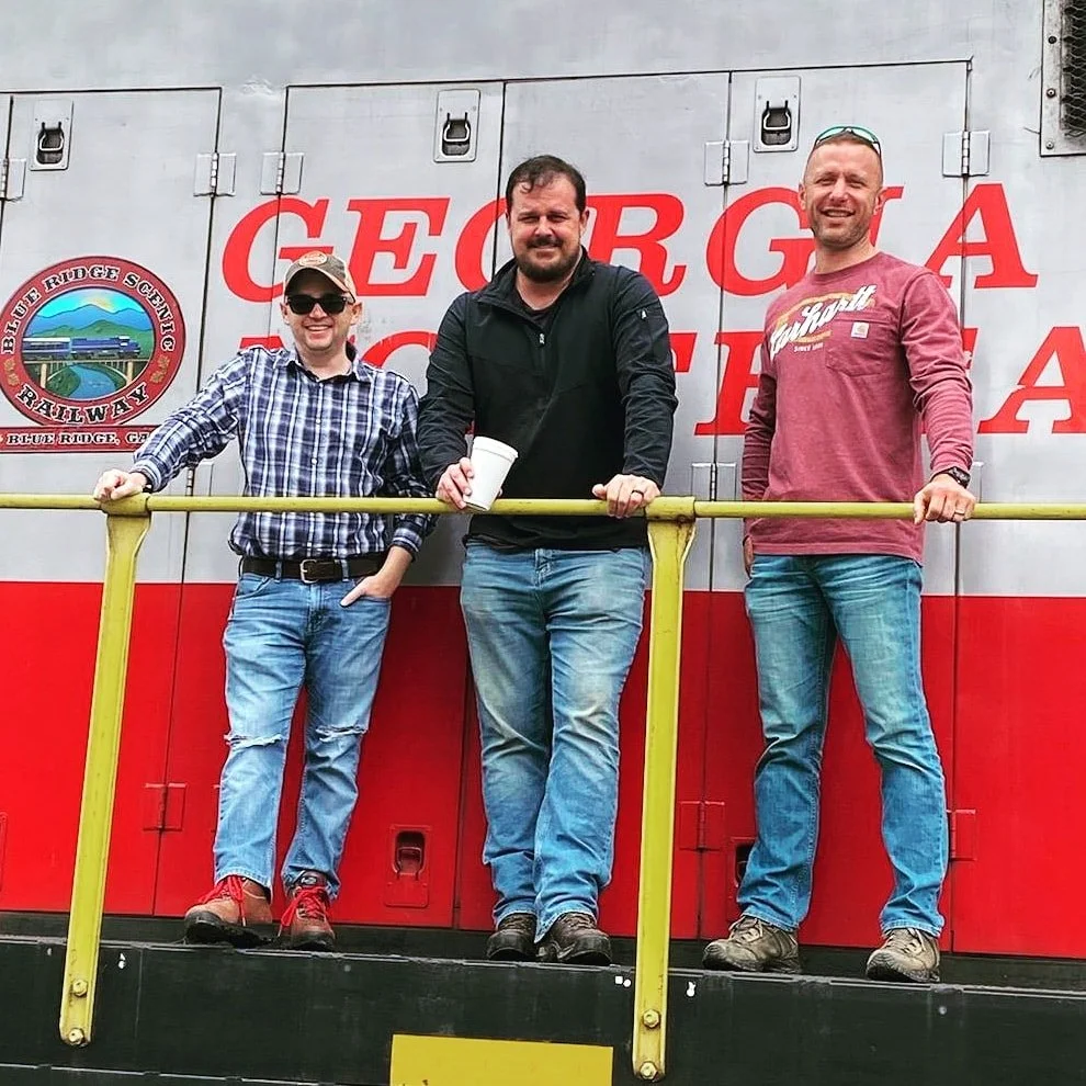 Three men standing on the platform of a train locomotive in North Georgia, posing for a photo during a railroad excursion in the Blue Ridge Mountains.