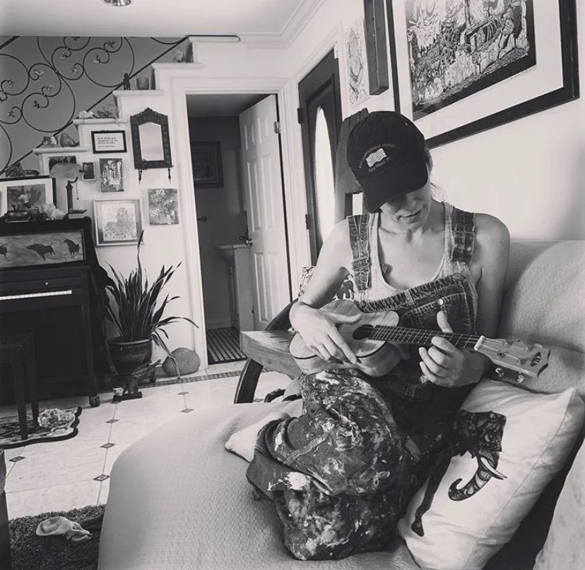 Woman sitting on a couch playing a ukulele in an art-filled living room.