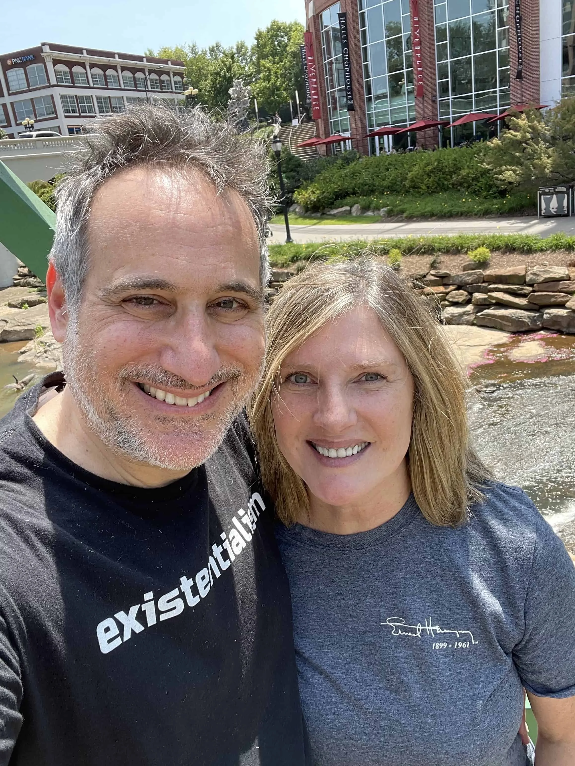 gray-hair man with a trimmed white beard stands in front of building with blonde haired smiling woman