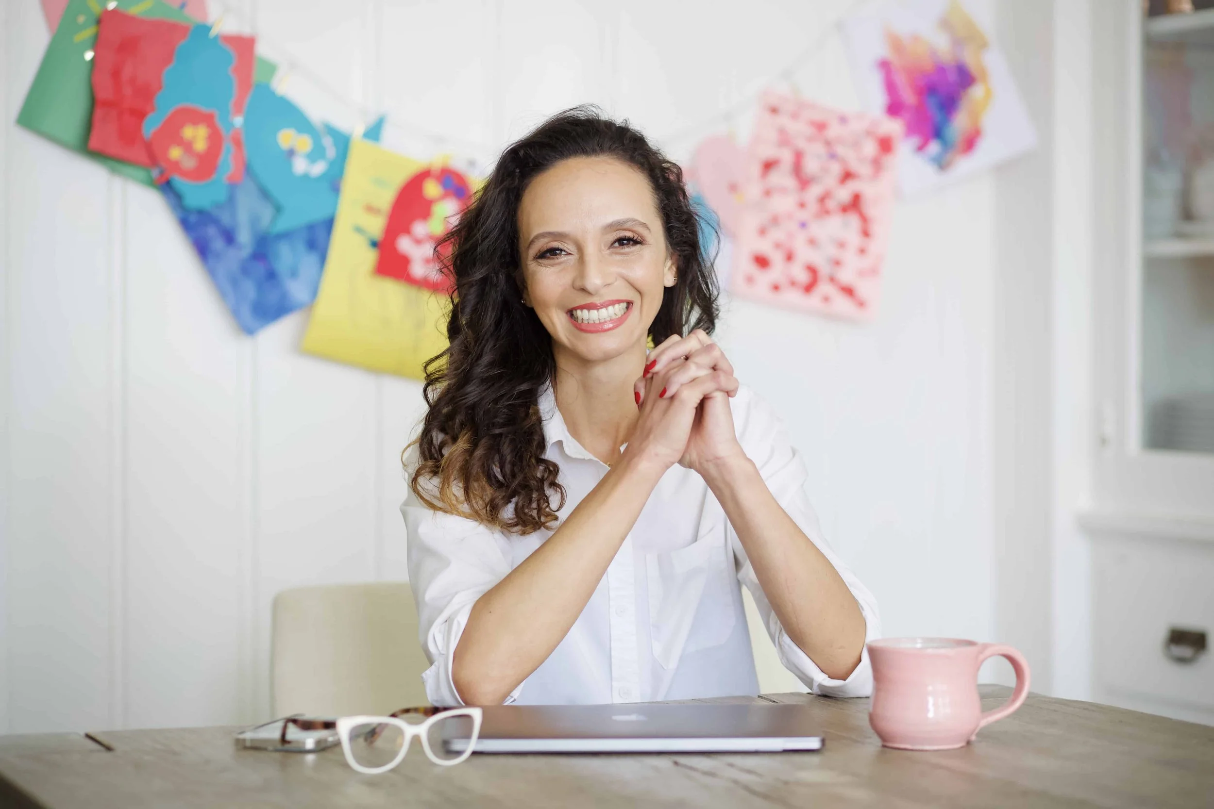 Oliveyah Fisch sits smiling at a wooden table with her hands clasped in front of her. A closed laptop, eyeglasses, and a pink mug rest on the table, with colorful artwork hanging on the white wall behind her.