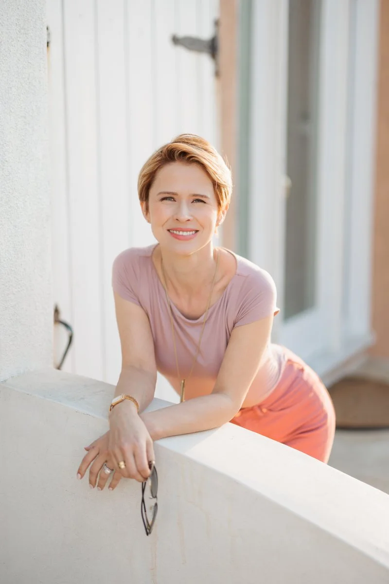 Woman with short blonde hair leans over a balcony with her arms crossed, smiling.