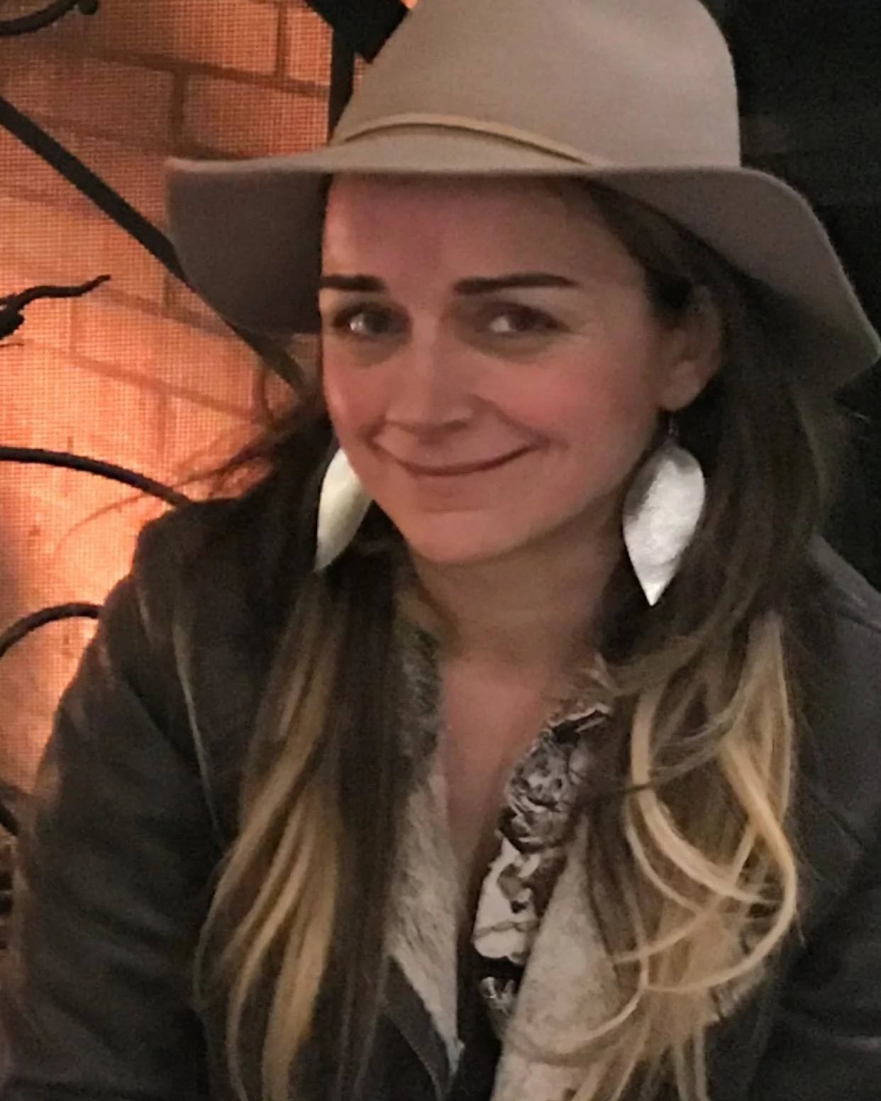 Woman smiling indoors while wearing a wide-brim hat and statement earrings.
