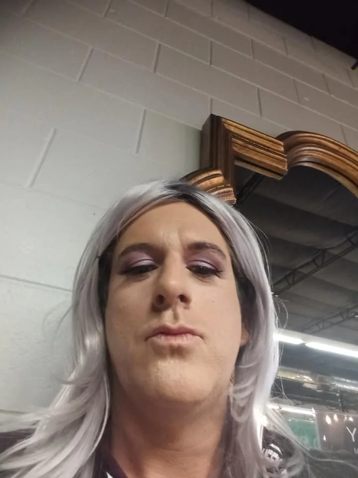Person with silver hair and purple eye makeup taking a selfie indoors near a decorative mirror and industrial-style ceiling.