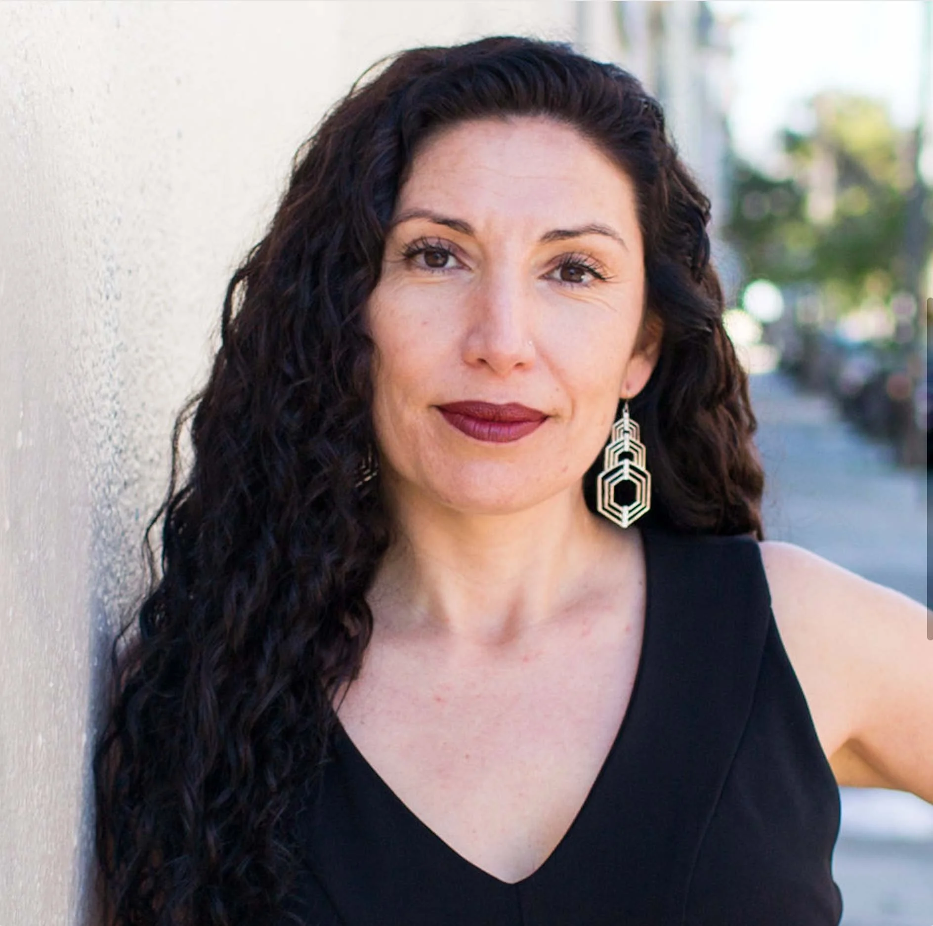 Maritza Schafer looks toward the camera in an outdoor portrait, wearing a black sleeveless top and geometric earrings.