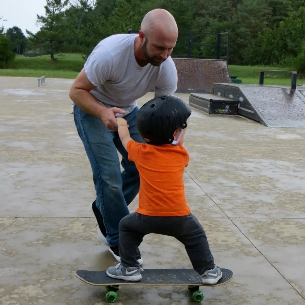 Man helping a young child balance on a skateboard at an outdoor skate park while holding the child's hands for support.