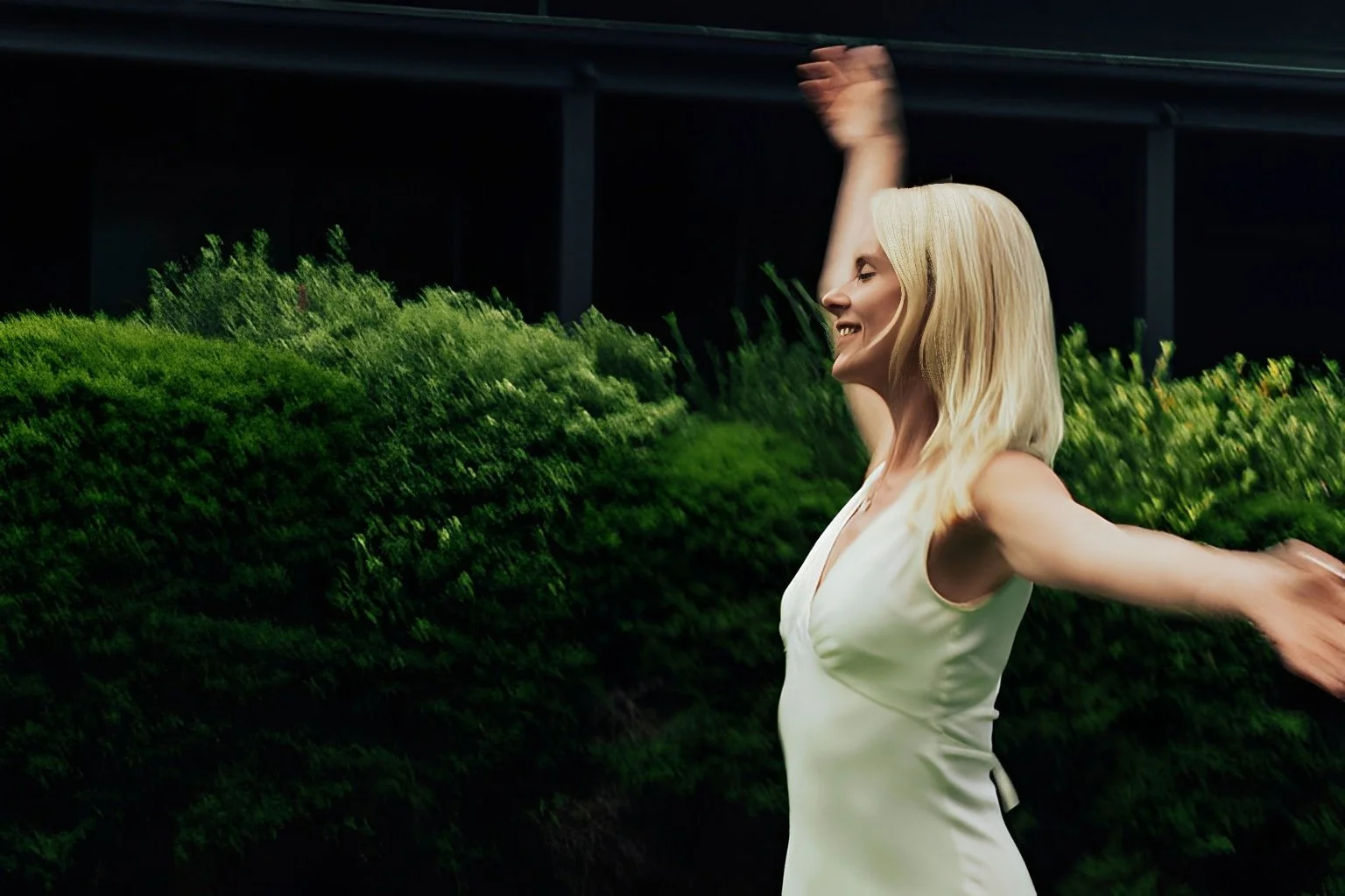 Blonde woman in a white dress standing outdoors at night with arms extended and eyes closed, appearing joyful and emotionally free.