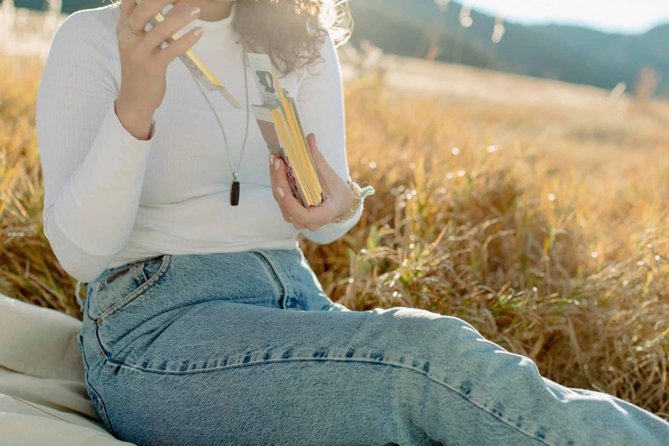 woman sits on grass in jeans and a white top shuffling a deck of tarot cards