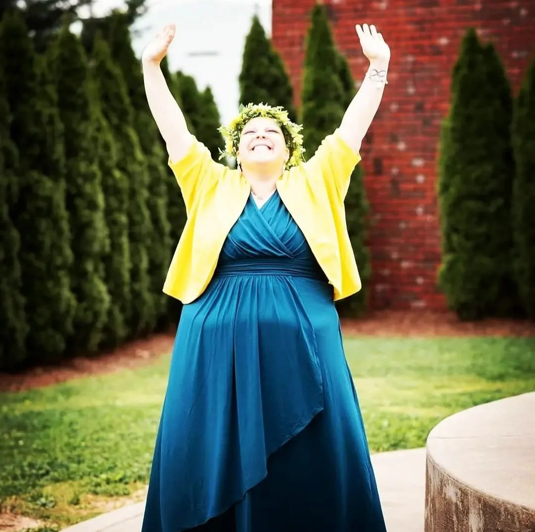 happy woman wears a lei on her head, blue dress, and yellow cardigan and throws her arms up in celebration