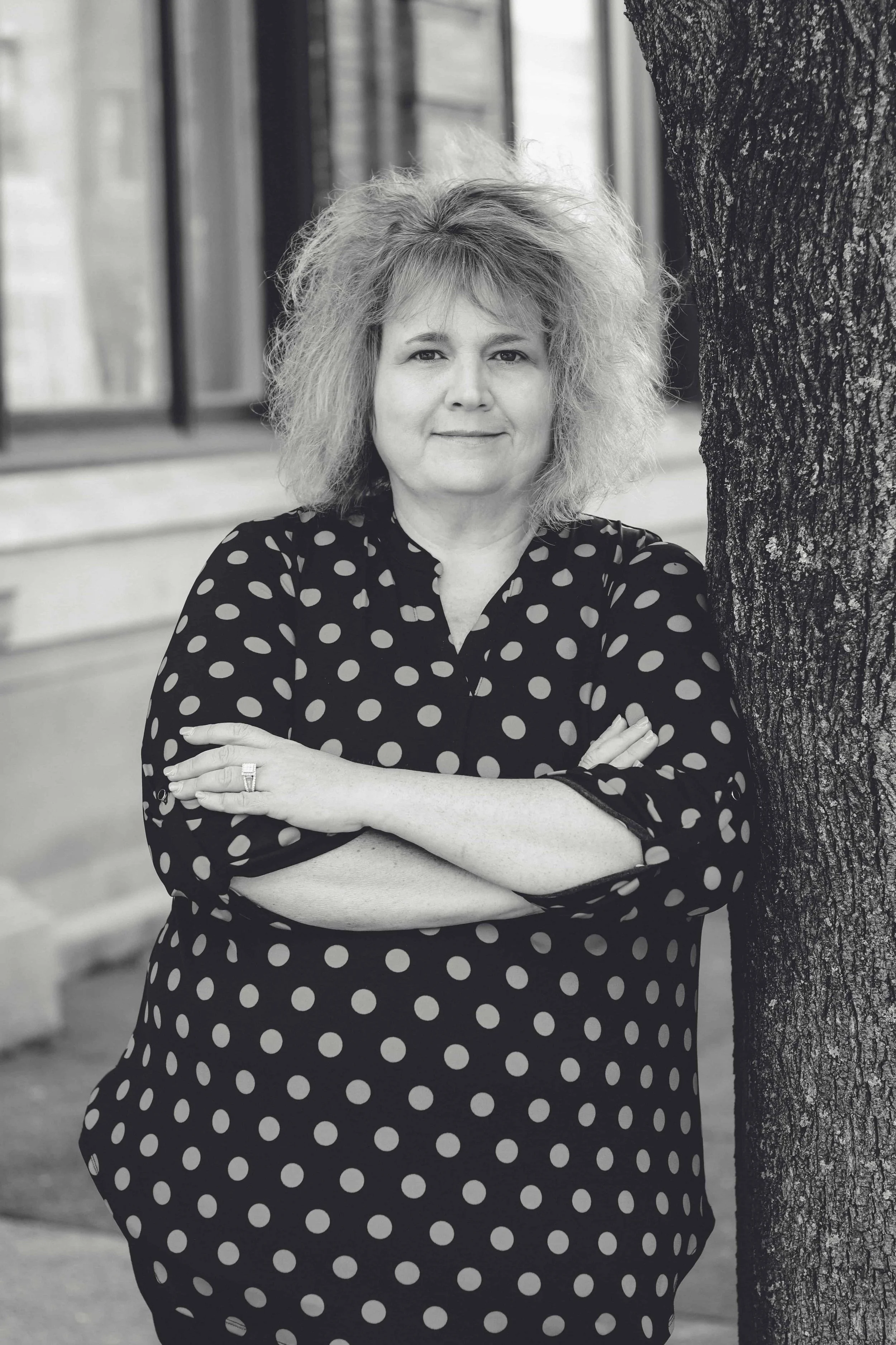 Black-and-white portrait of a woman leaning against a tree with her arms crossed and a slight smile on her face.