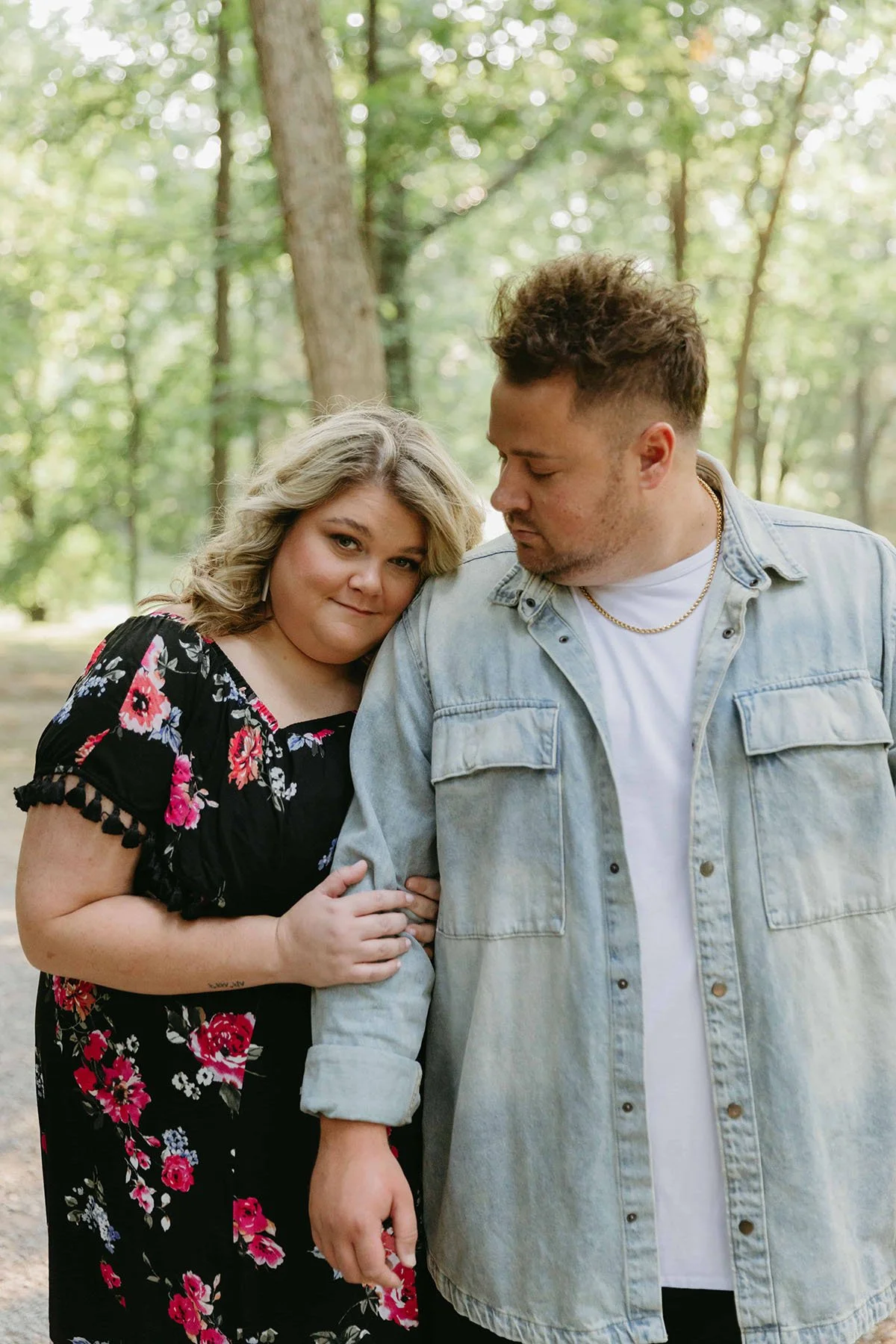 Couple posing together outdoors in a wooded area, with the woman resting her head on the man’s shoulder while he looks at her.