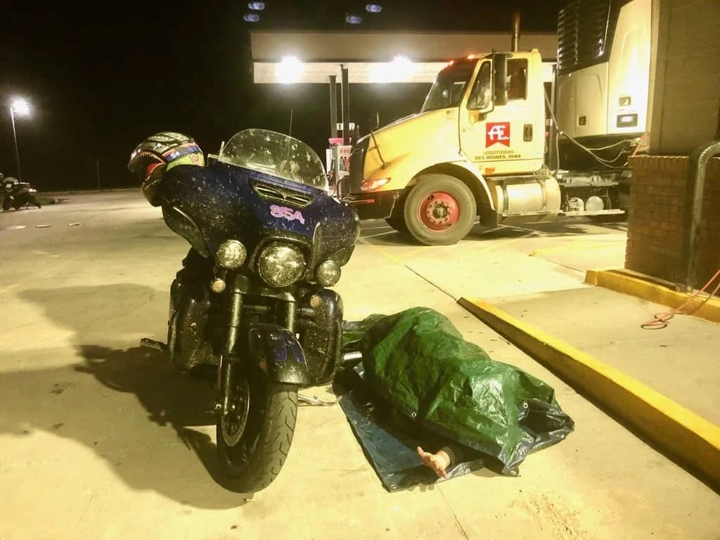 Motorcycle rider laying down to sleep under tarp next to motorcycle.