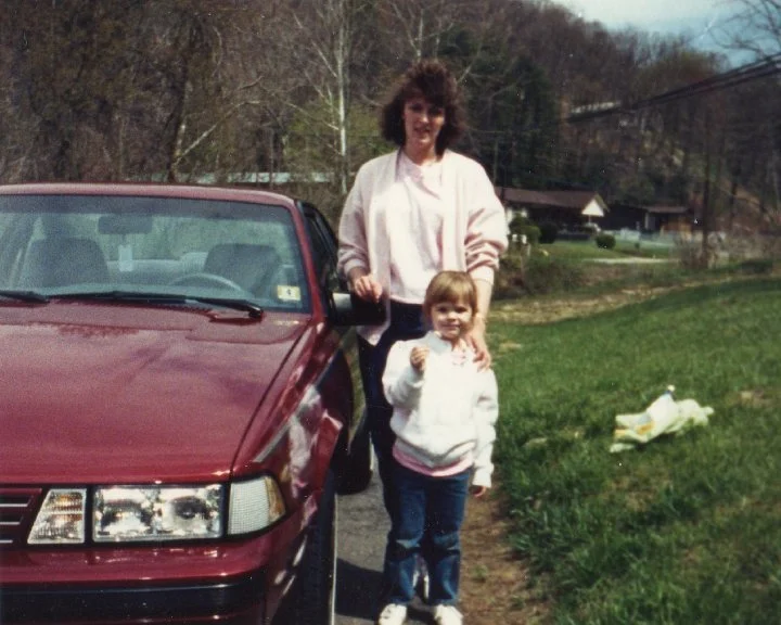Adult mother and child standing beside a red car in a grassy rural area.