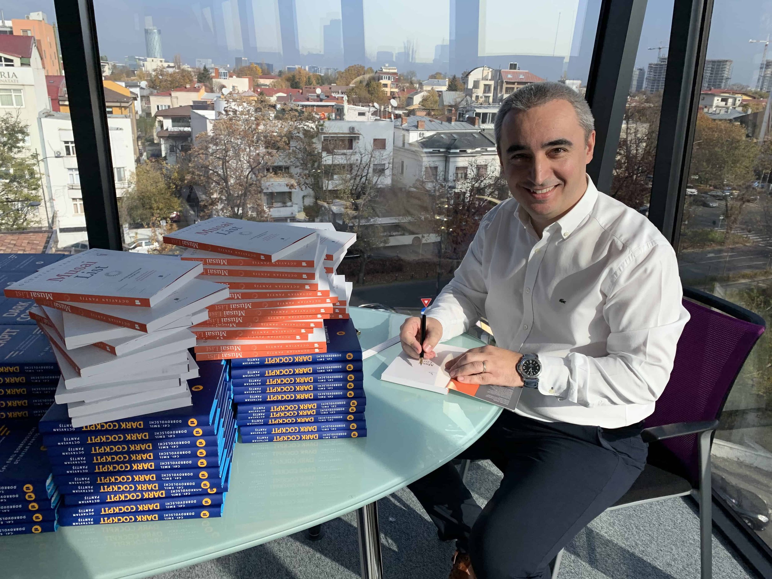 Smiling man sits at table autographing a stack of books.
