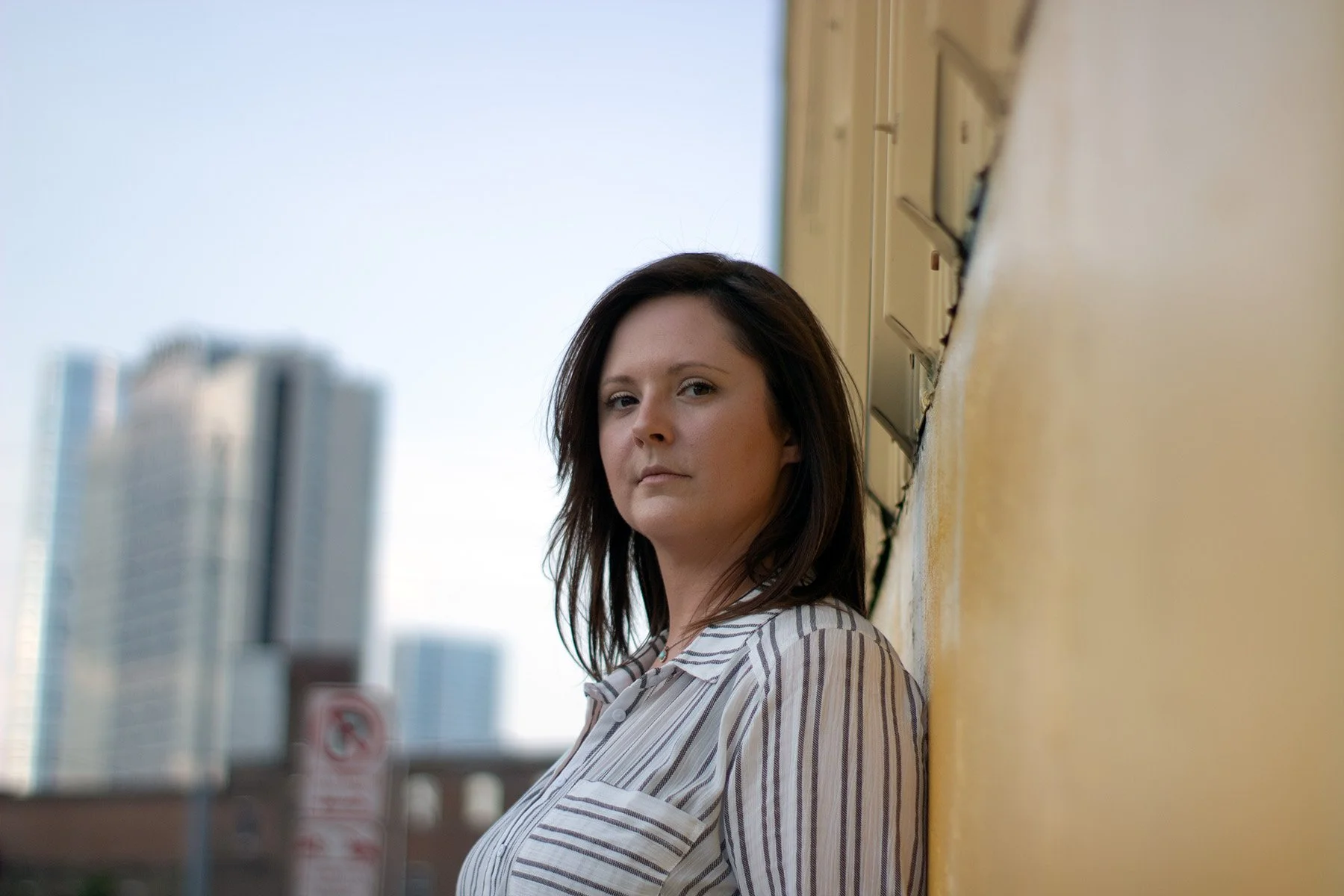 A woman with dark shoulder-length hair leans against a yellow wall outdoors, looking directly at the camera with a city skyline blurred in the background.