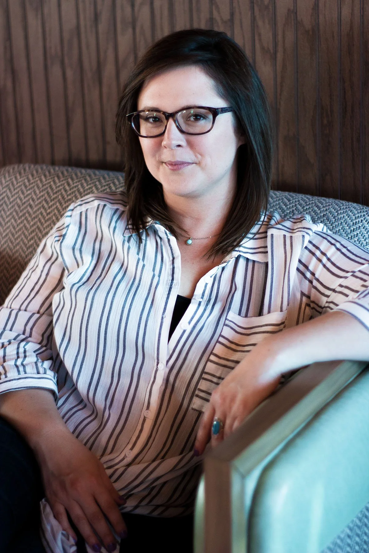 A woman with dark hair and glasses sits on a couch, resting her arm on the armrest and smiling gently at the camera.