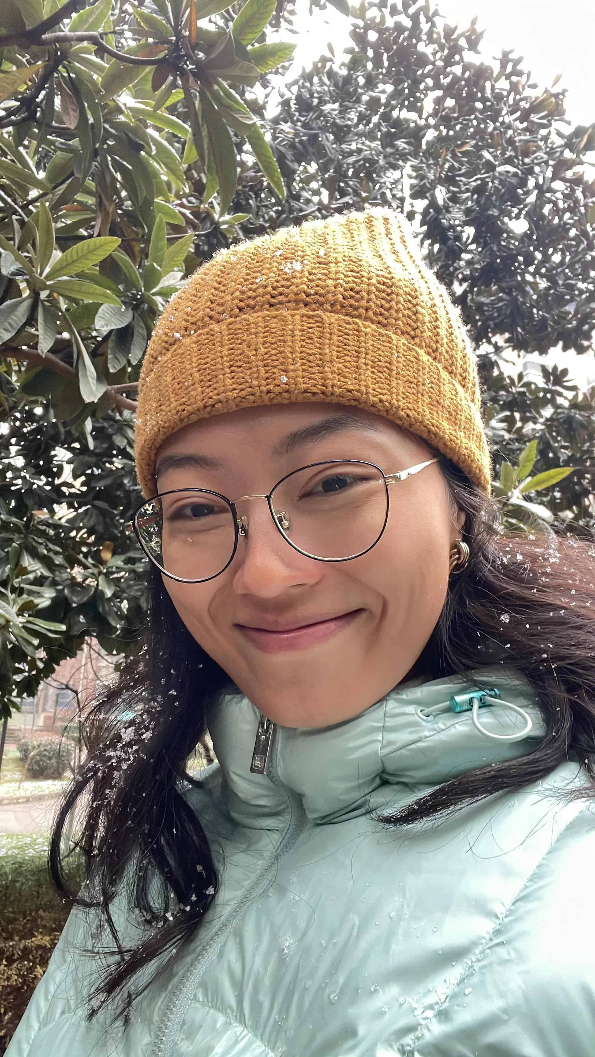 Asian woman wearing a light green winter jacket smiling with snow on her hair