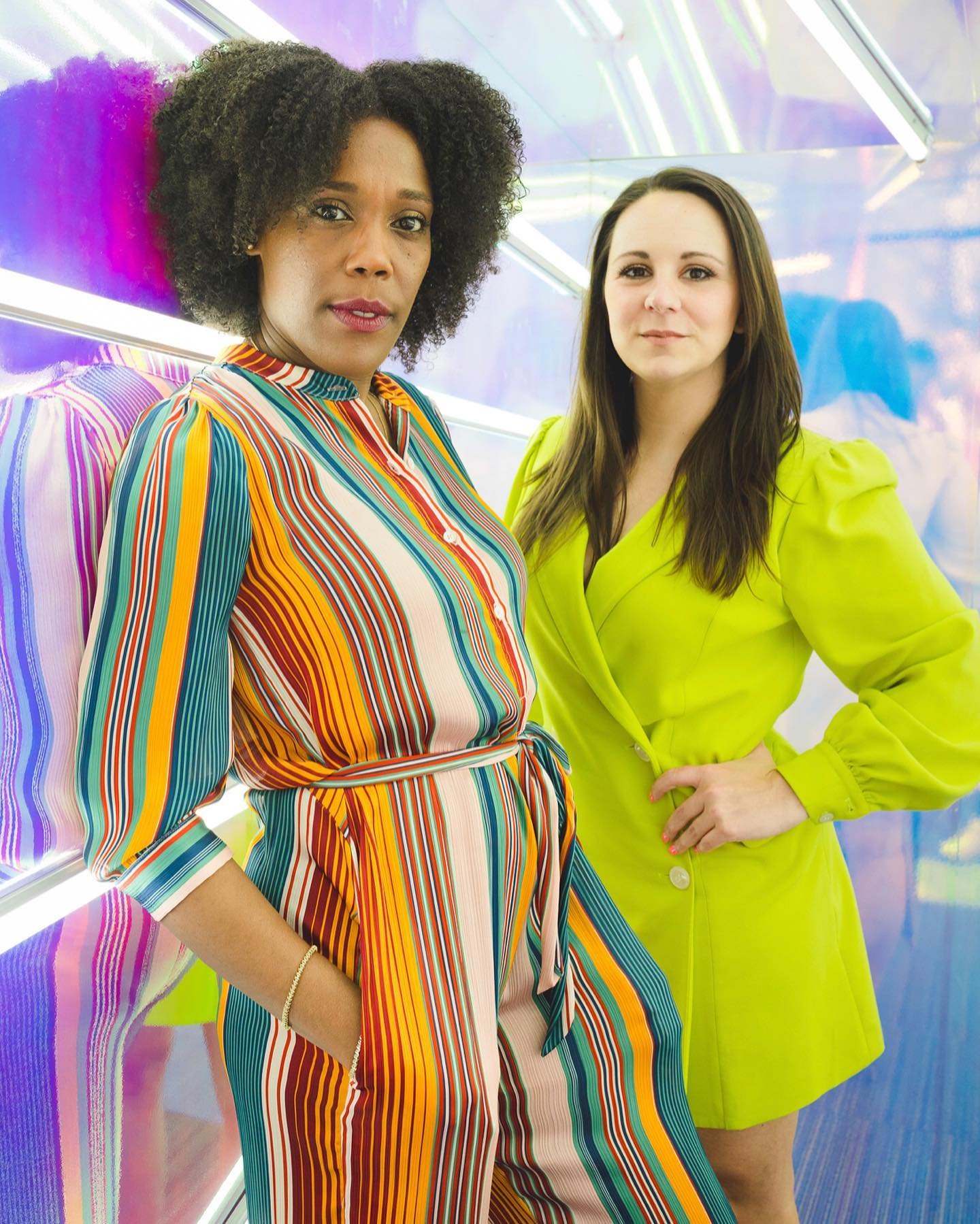 Two female founders posing in front of a colorful reflective wall, wearing a lime green dress and a pinstriped jumpsuit.