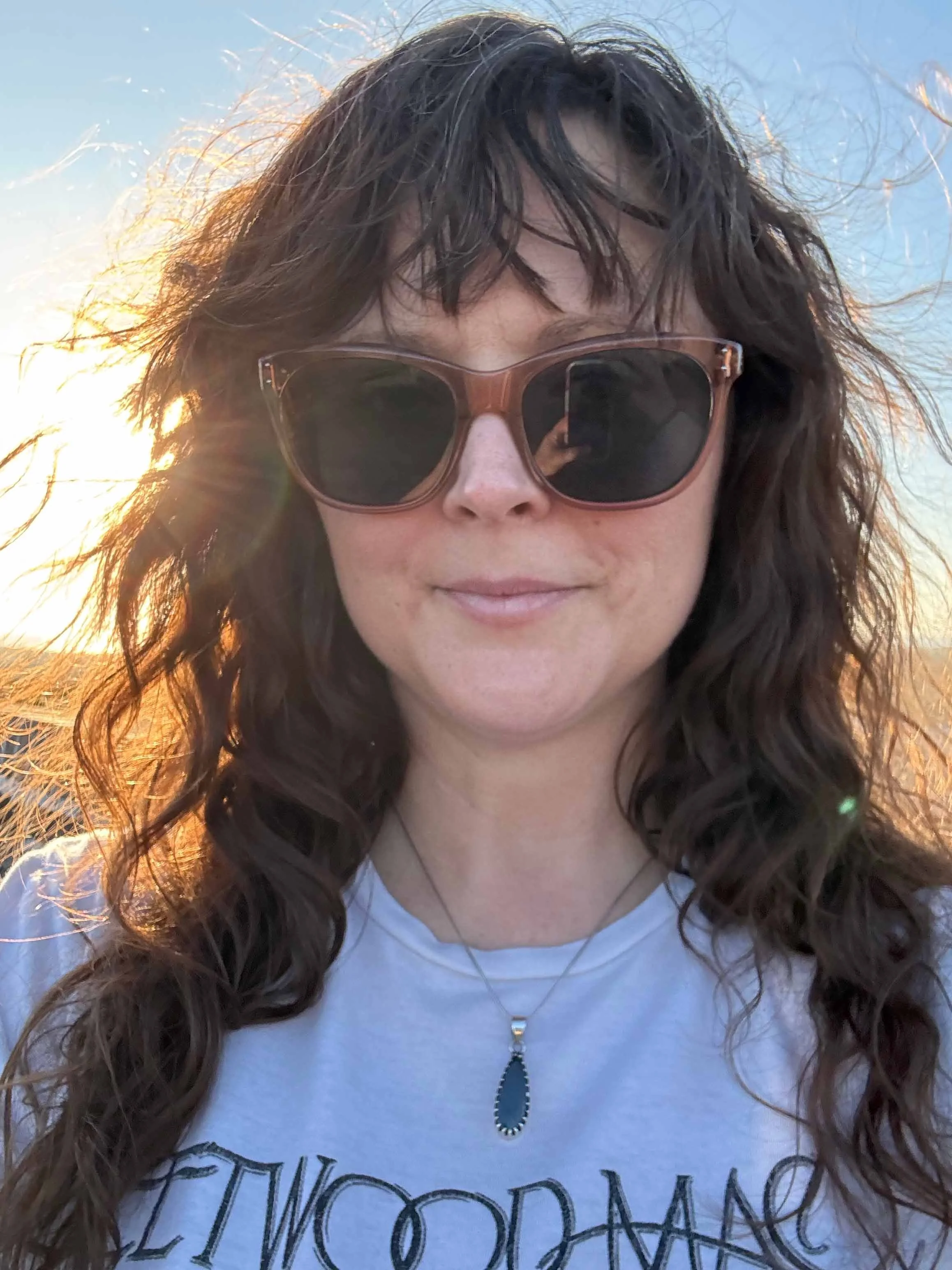 Woman outdoors wearing sunglasses and a polished teardrop-shaped hematite pendant on a silver chain, with sunlight glowing through her dark wavy hair.