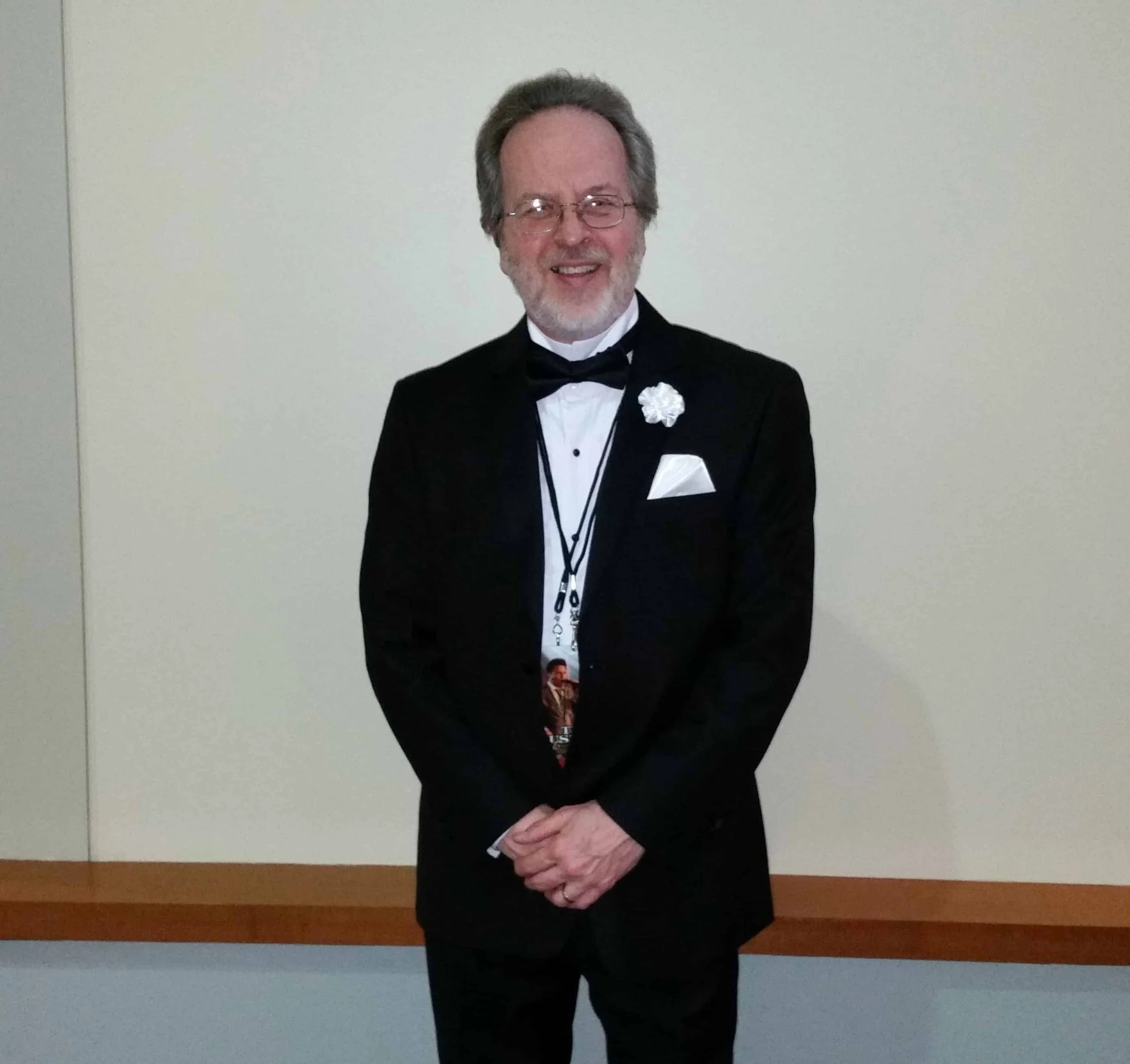 Smiling man wearing glasses, a tuxedo, and a backstage pass stands with his hands folded in front of him.