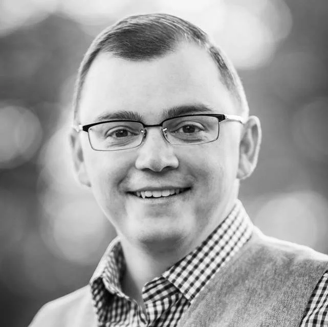 Smiling man in glasses wearing a checkered shirt and sweater vest.