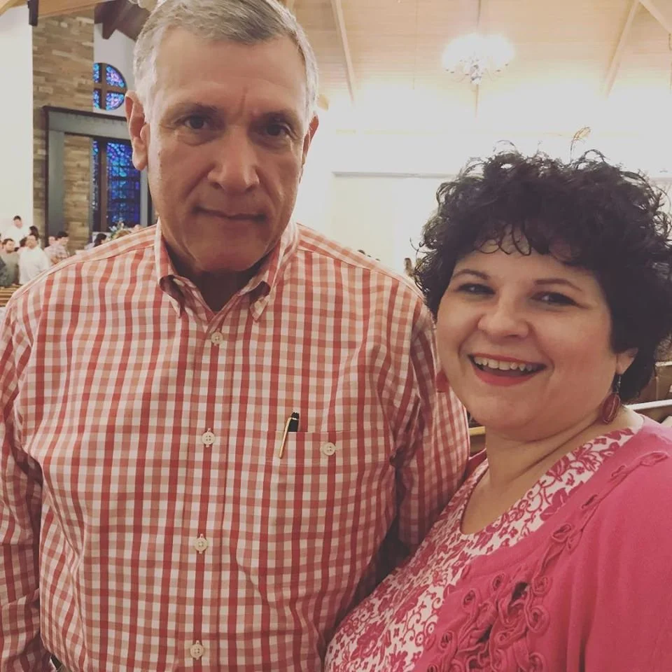 A smiling couple standing together inside a church with stained glass windows in the background.