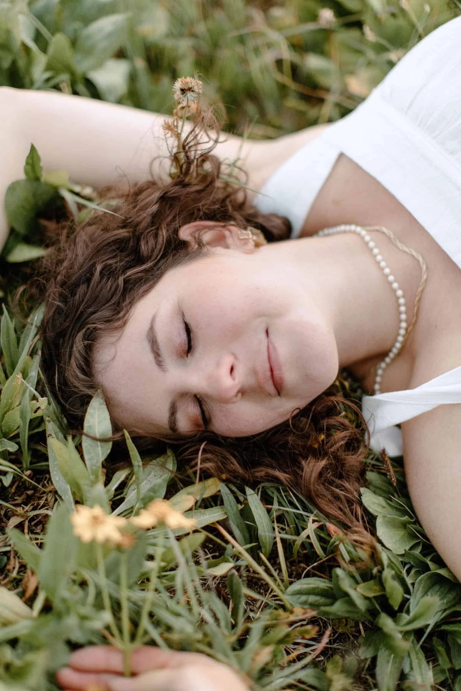 brunette woman in a white tank top lays on the grass with her eyes closed and a peaceful expression