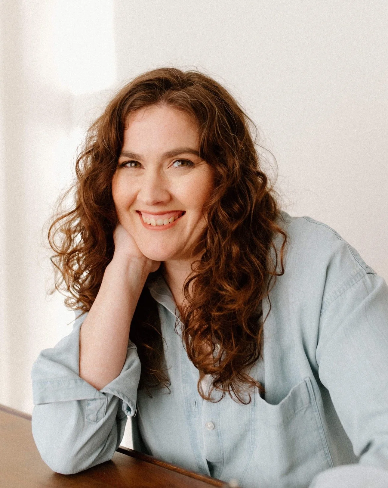Curly haired woman smiles for a portrait with her head resting on her hand.