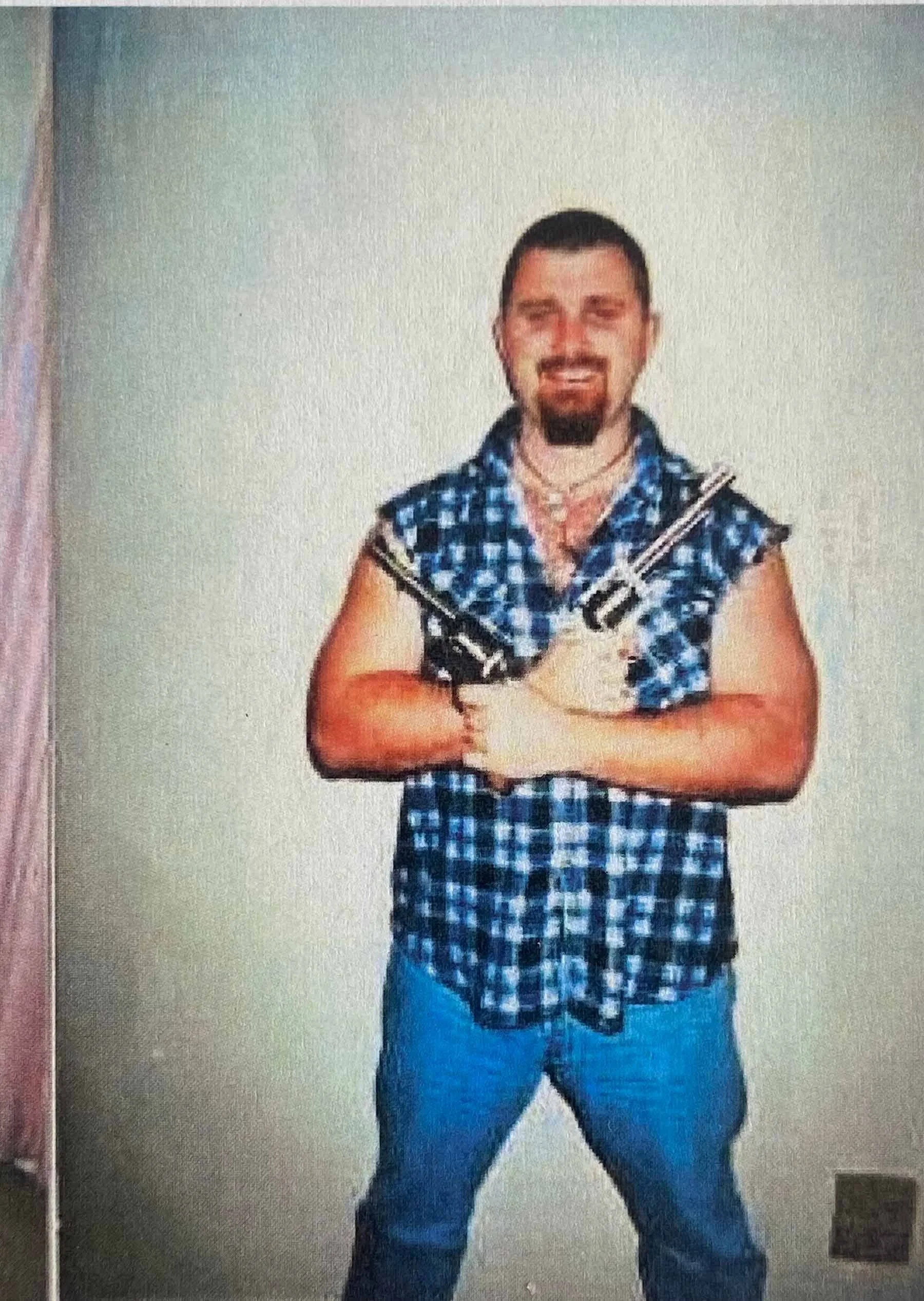 An older photo of Todd Maguire during his undercover police years, standing against a plain wall in a sleeveless plaid shirt and jeans while holding two handguns across his chest.