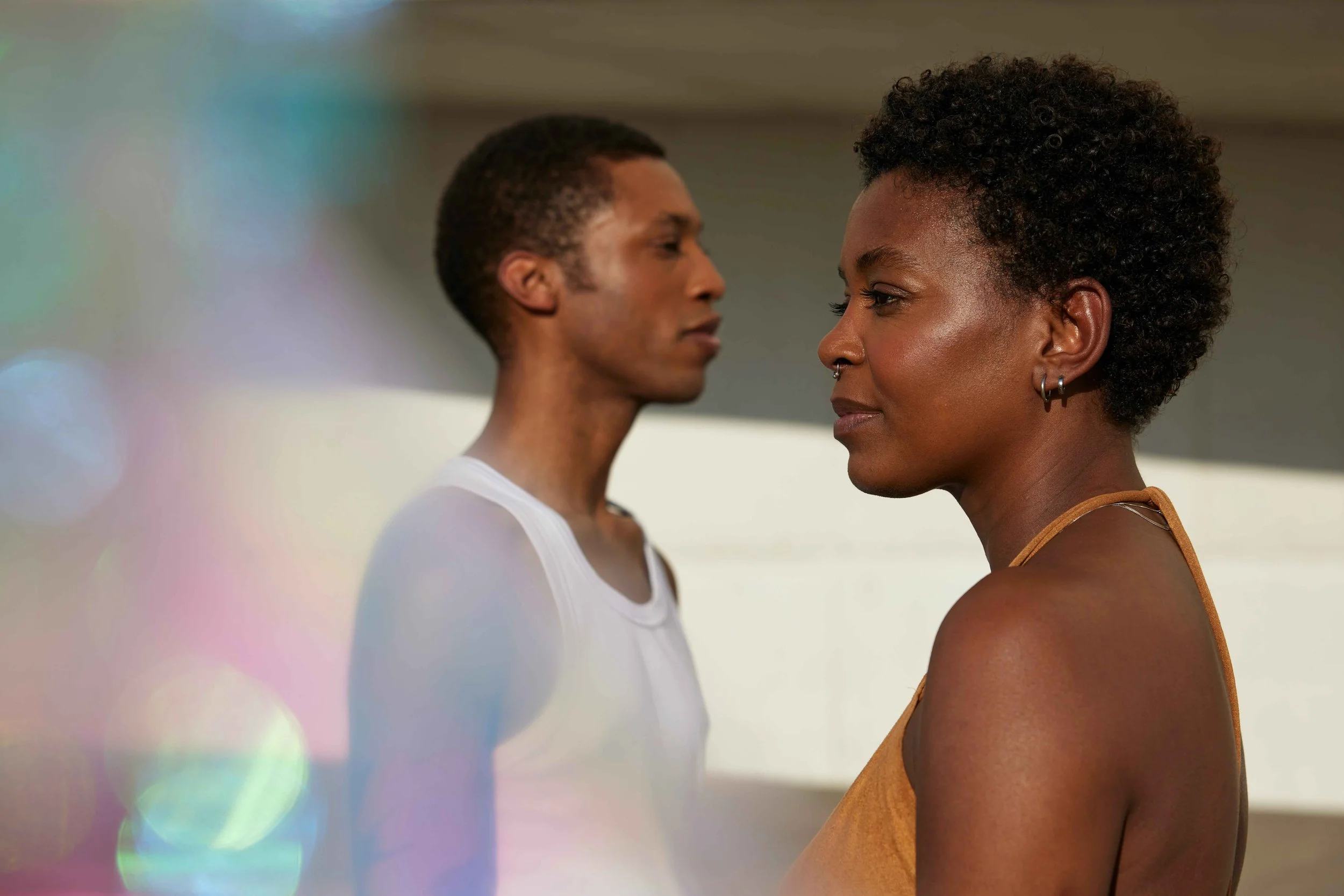 Two people standing side by side outdoors, one in focus in the foreground and the other softly blurred in the background, both looking off into the distance.