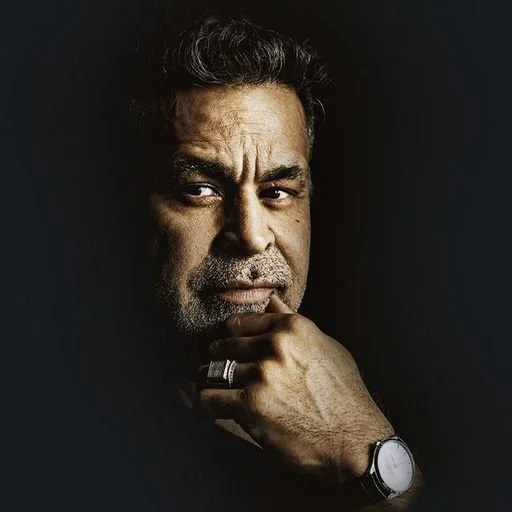 Dramatic portrait of a man resting his hand on his chin and looking thoughtfully to the side against a dark background.