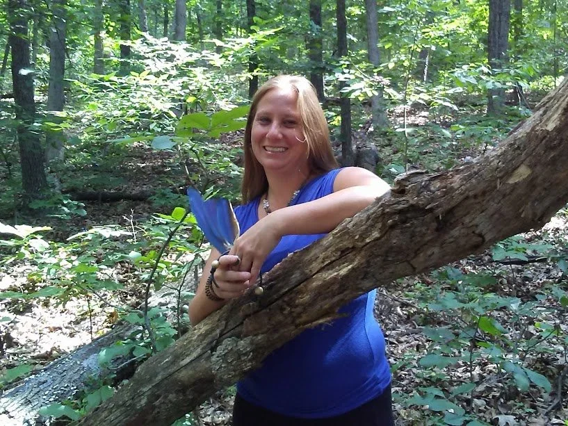 A woman in a blue shirt smiles in a green forest while standing behind a fallen tree and holding feathers.
