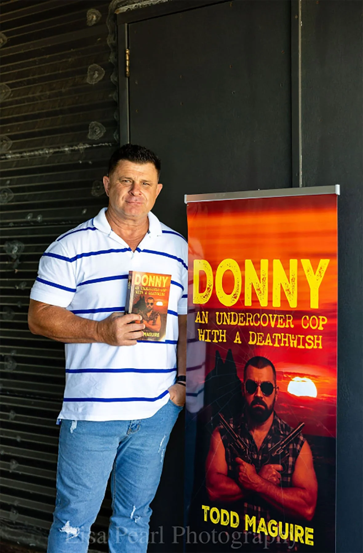 Todd Maguire stands beside a large banner for Donny: An Undercover Cop with a Deathwish while holding a copy of the book.