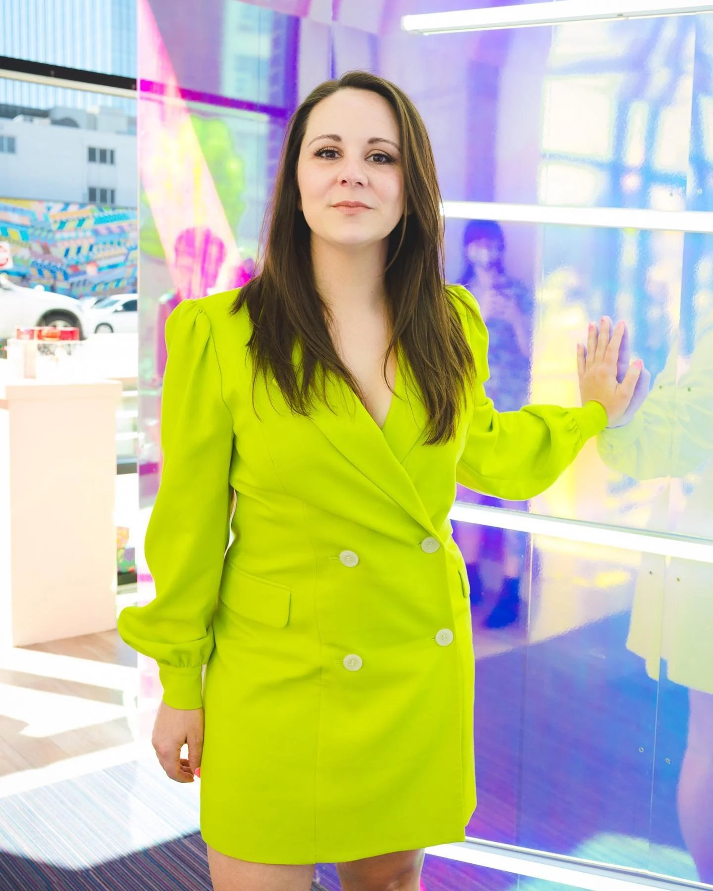 Woman in a bright lime green dress posing with one hand against a colorful reflective wall.