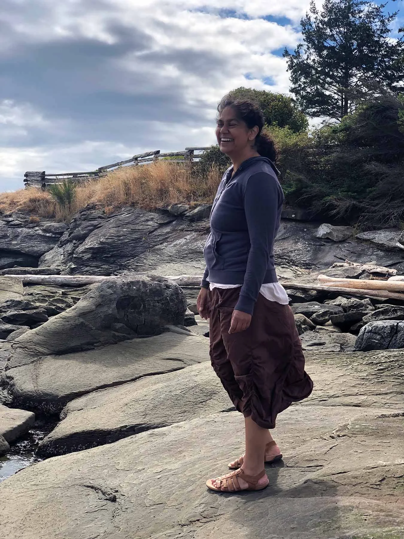 Natasha Ramlall smiling while standing on a rocky shoreline beneath a cloudy sky, with trees and driftwood in the background.