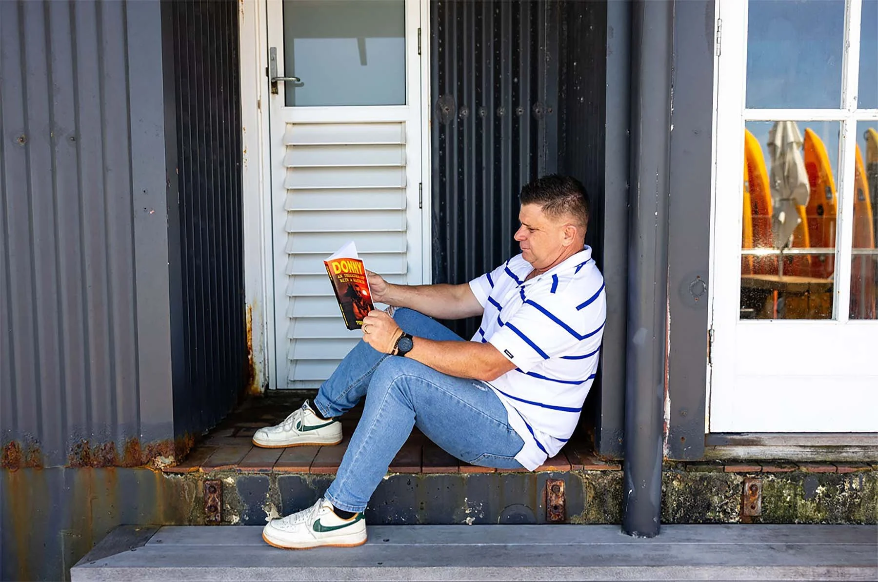 Todd Maguire sits on a doorstep reading a copy of his book, Donny: An Undercover Cop with a Deathwish, outside a dark corrugated building.