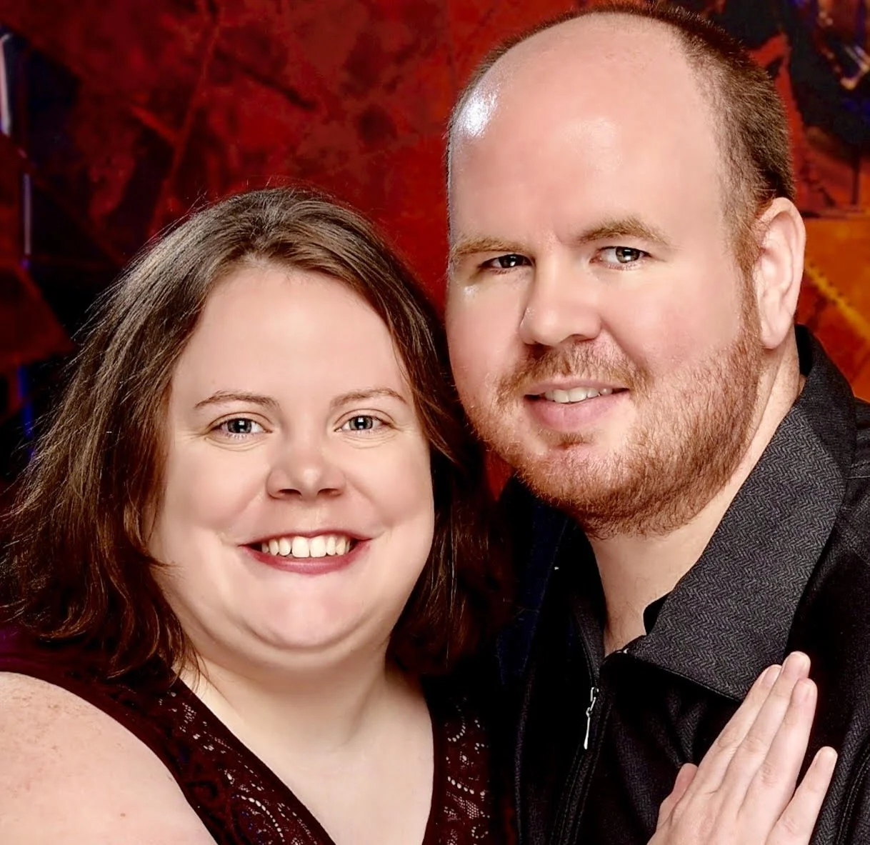 A smiling couple posing together for a portrait against a warm red background.