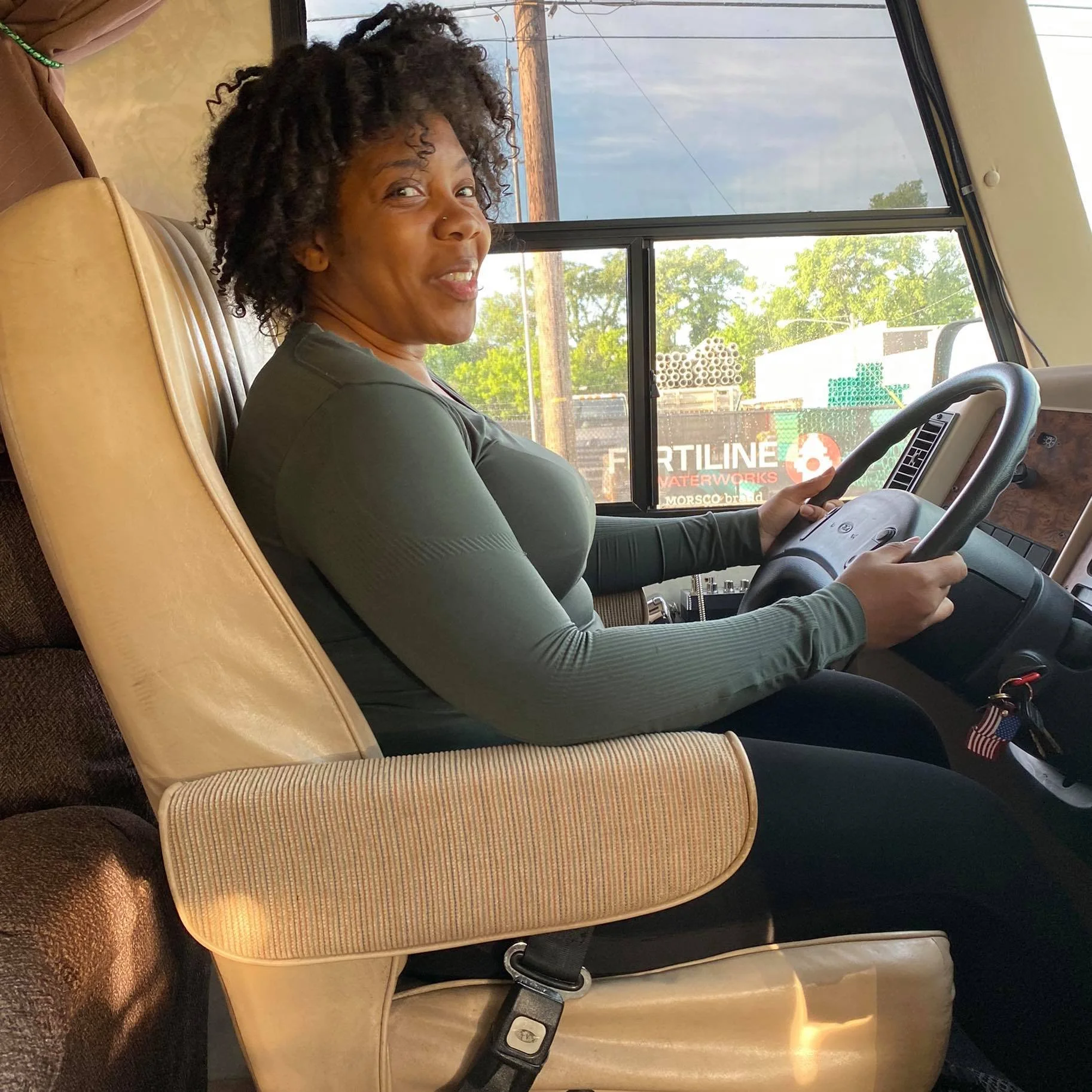 Woman driving an RV while smiling at the camera, wearing athletic clothing and seated behind the steering wheel in daylight.