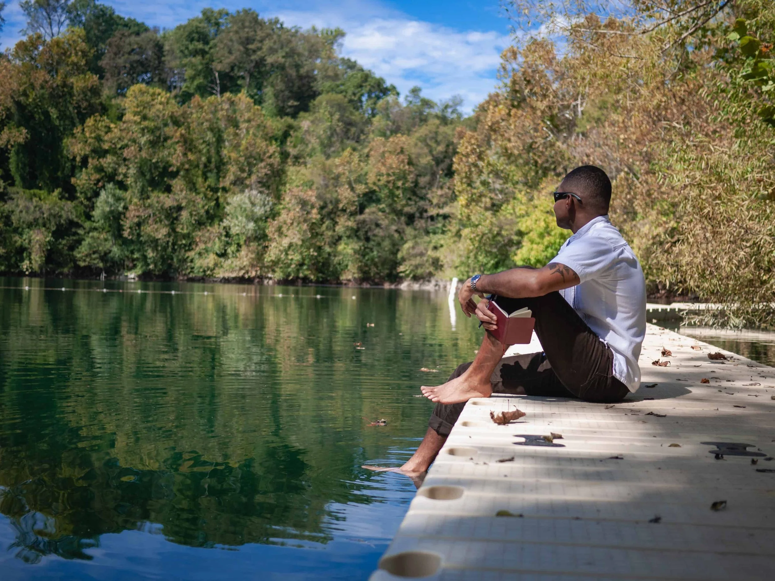 Ty Briggs sits barefoot on a dock beside a calm green lake, holding a journal while looking out toward dense trees and blue sky in a peaceful natural setting.