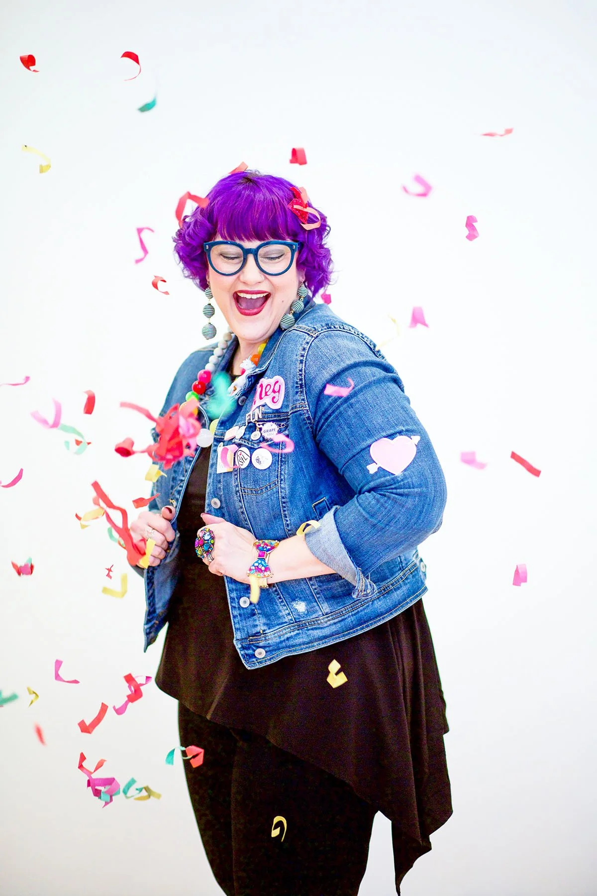 Person with curly purple hair, colorful accessories, and a denim jacket smiles as colorful confetti falls around them against a white studio background.
