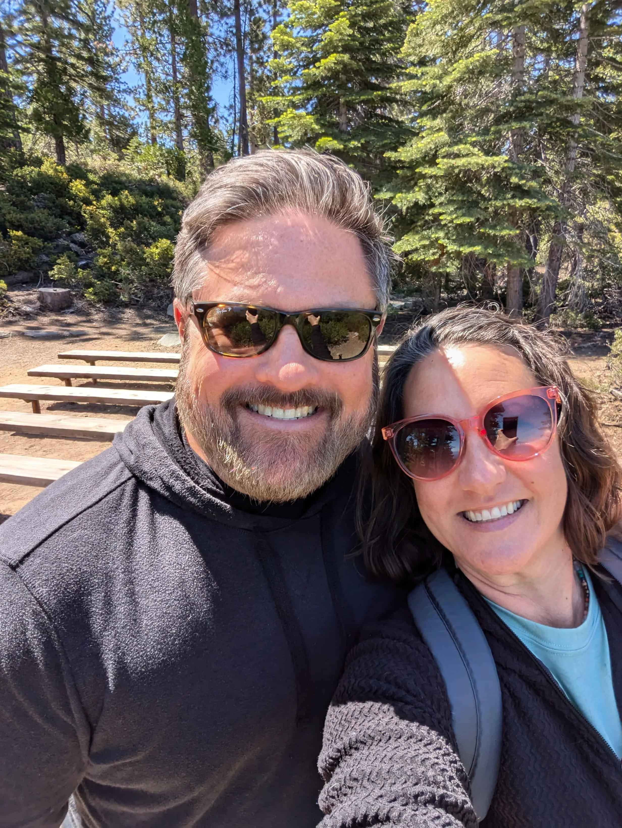 Couple standing close together, smiling for a selfie in an outdoor amphitheater with tiered seating visible behind them.