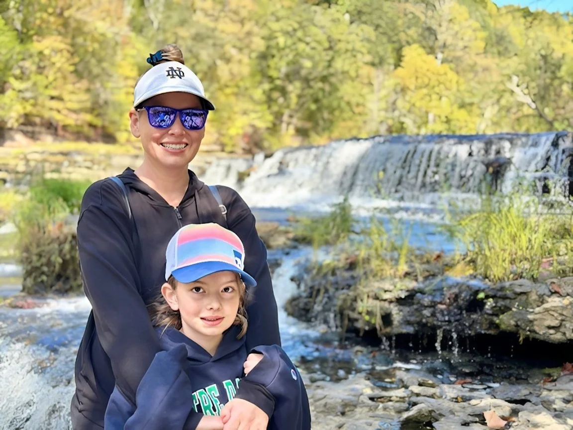 Woman in hat with sunglasses hugging a little girl in a hat and hoodie in front of a waterfall.