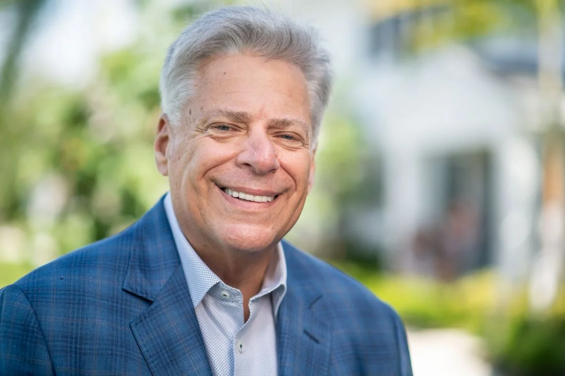 Smiling man in a blue suit jacket posing outdoors with a blurred residential background.