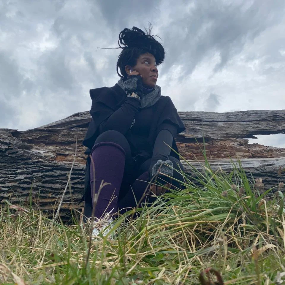Woman sitting outdoors on grass near a large fallen tree under cloudy skies, wearing dark layered clothing and looking thoughtfully into the distance.