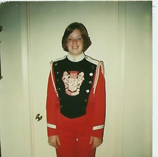 Childhood photo of teenage girl standing indoors in a red marching band uniform.
