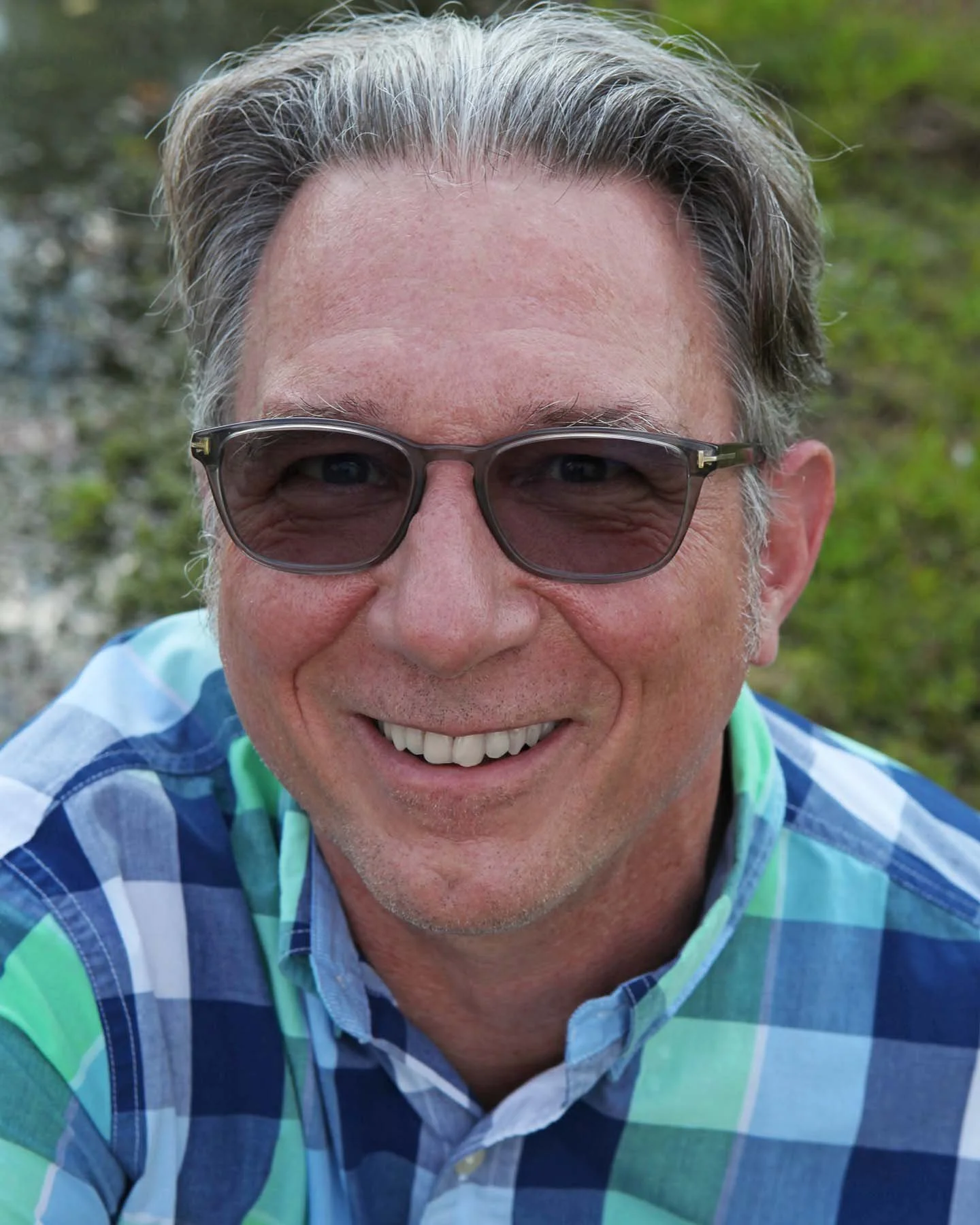 Smiling man with gray hair and glasses wearing a blue and green plaid shirt outdoors near a stream.