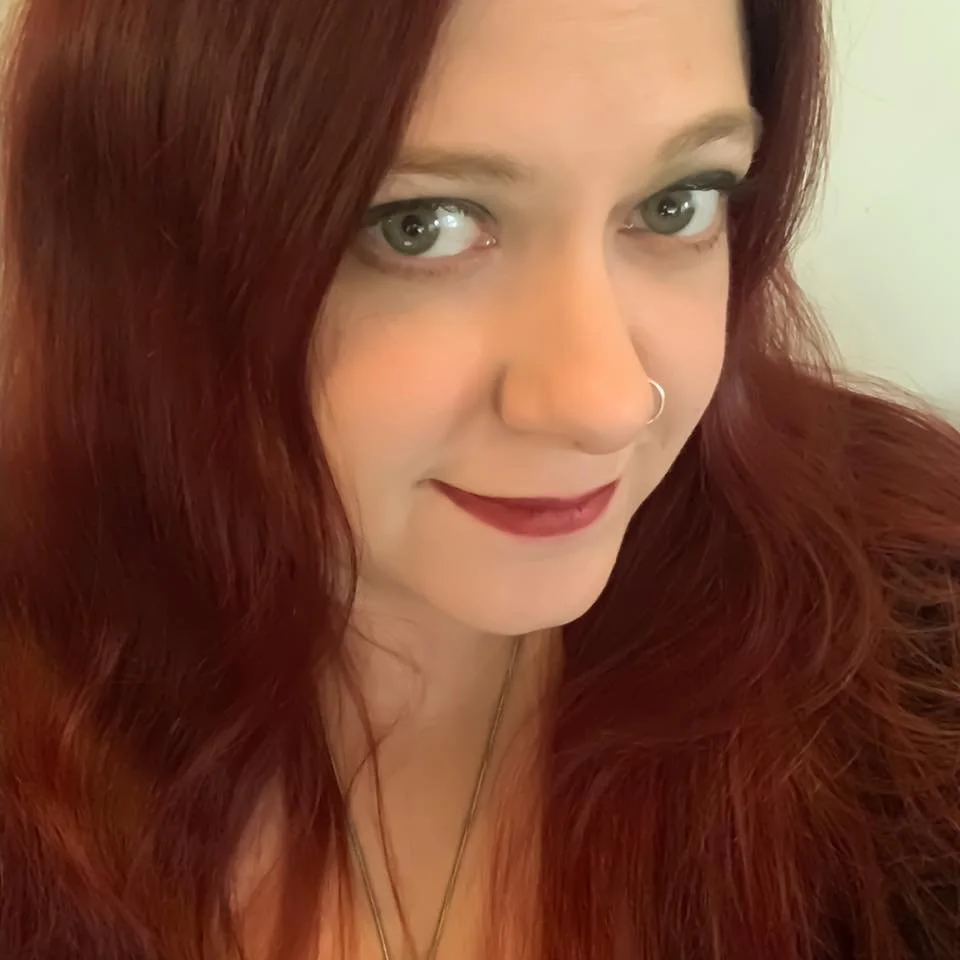 Portrait of a woman with long red hair, a nose ring, and burgundy lipstick looking off camera.
