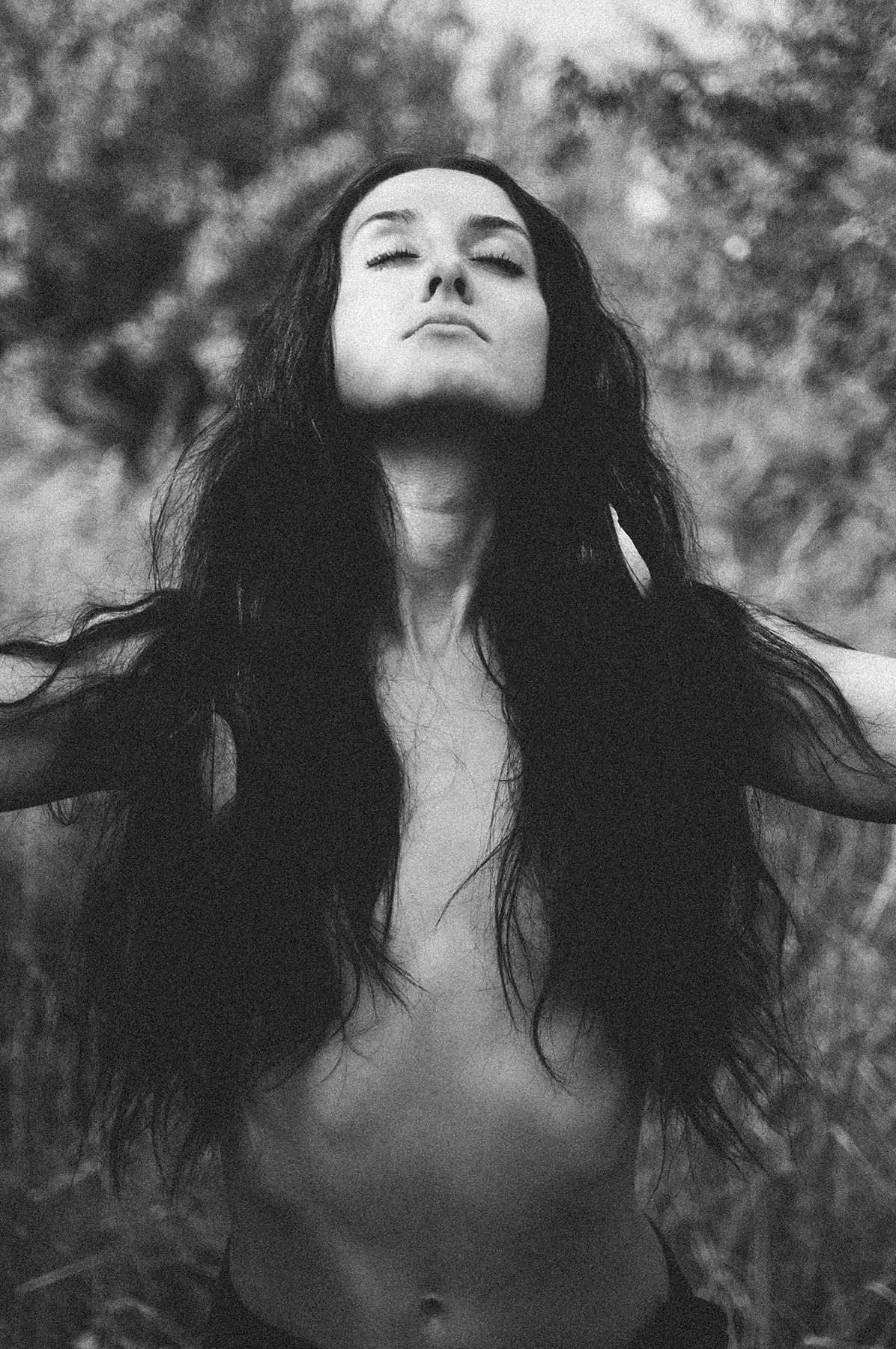Black-and-white portrait of a woman with long dark hair standing outdoors with her head tilted upward and eyes closed, surrounded by a softly blurred natural background.