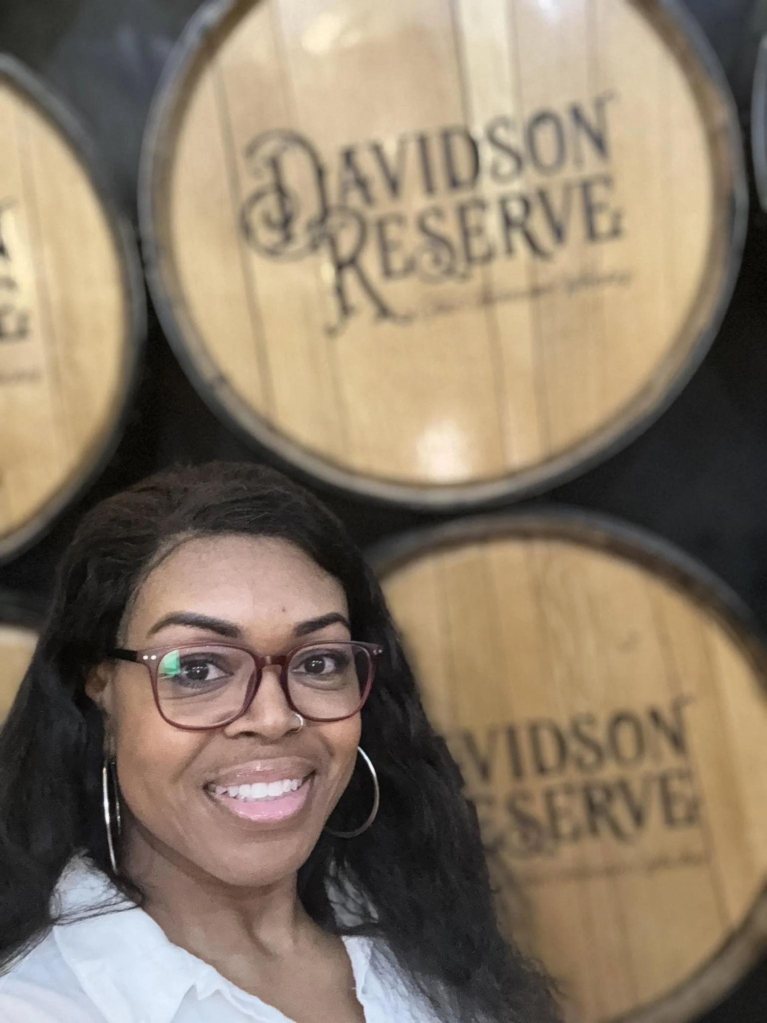 Smiling Black woman wearing red glasses, silver hoops, and white shirt stands in front of bourbon barrels