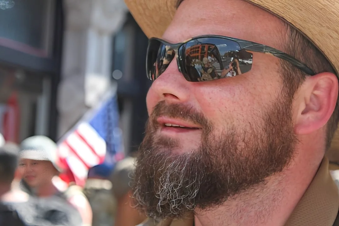 Close-up of a bearded man wearing sunglasses and a tan hat at an outdoor event with an American flag behind him.